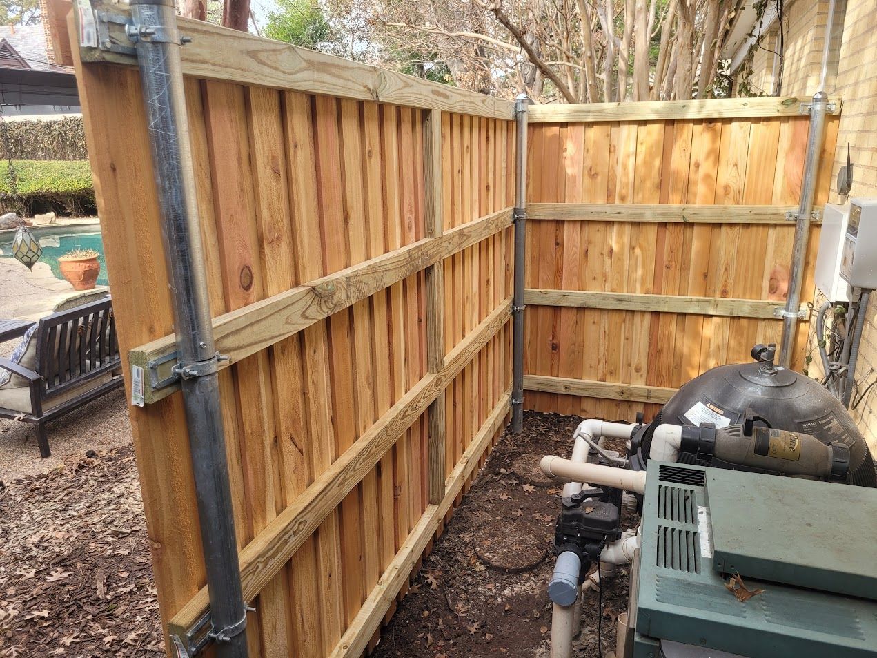 Wooden backyard fence around equipment and pipes, with a black tank and green machine beside gravel.
