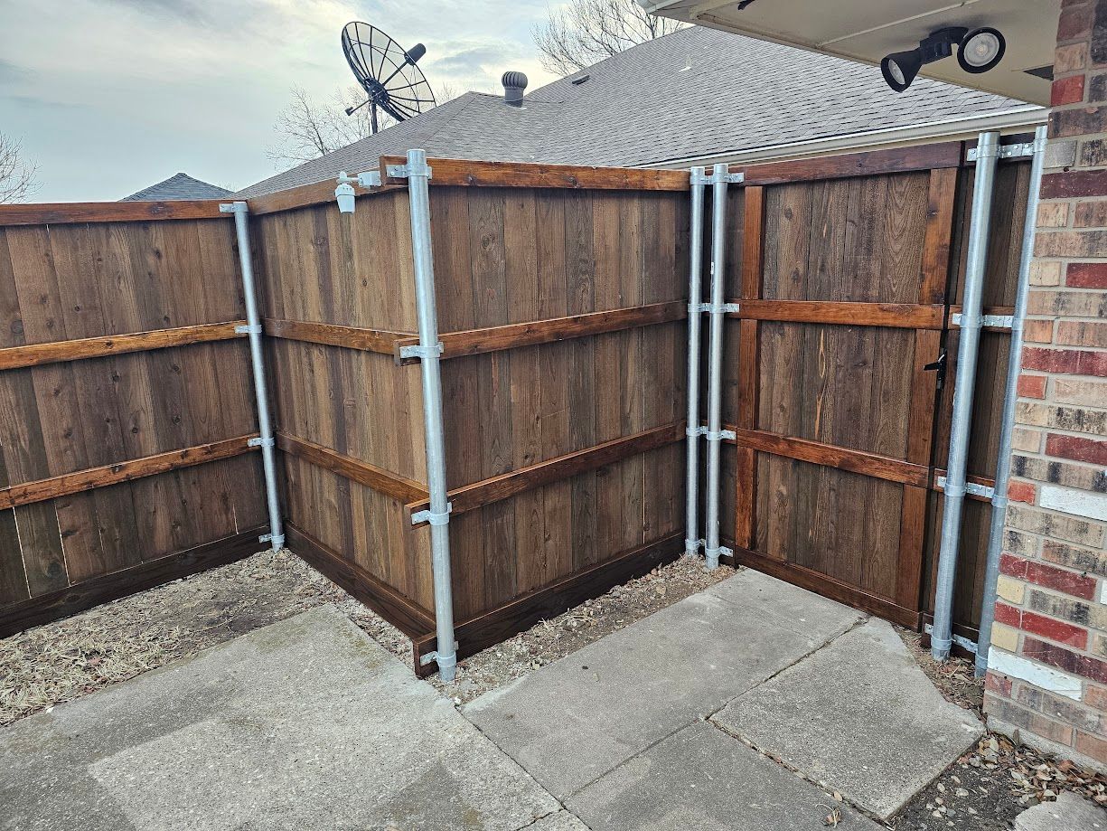 Brown wooden fence panels and metal posts around a paved backyard corner beside a brick wall