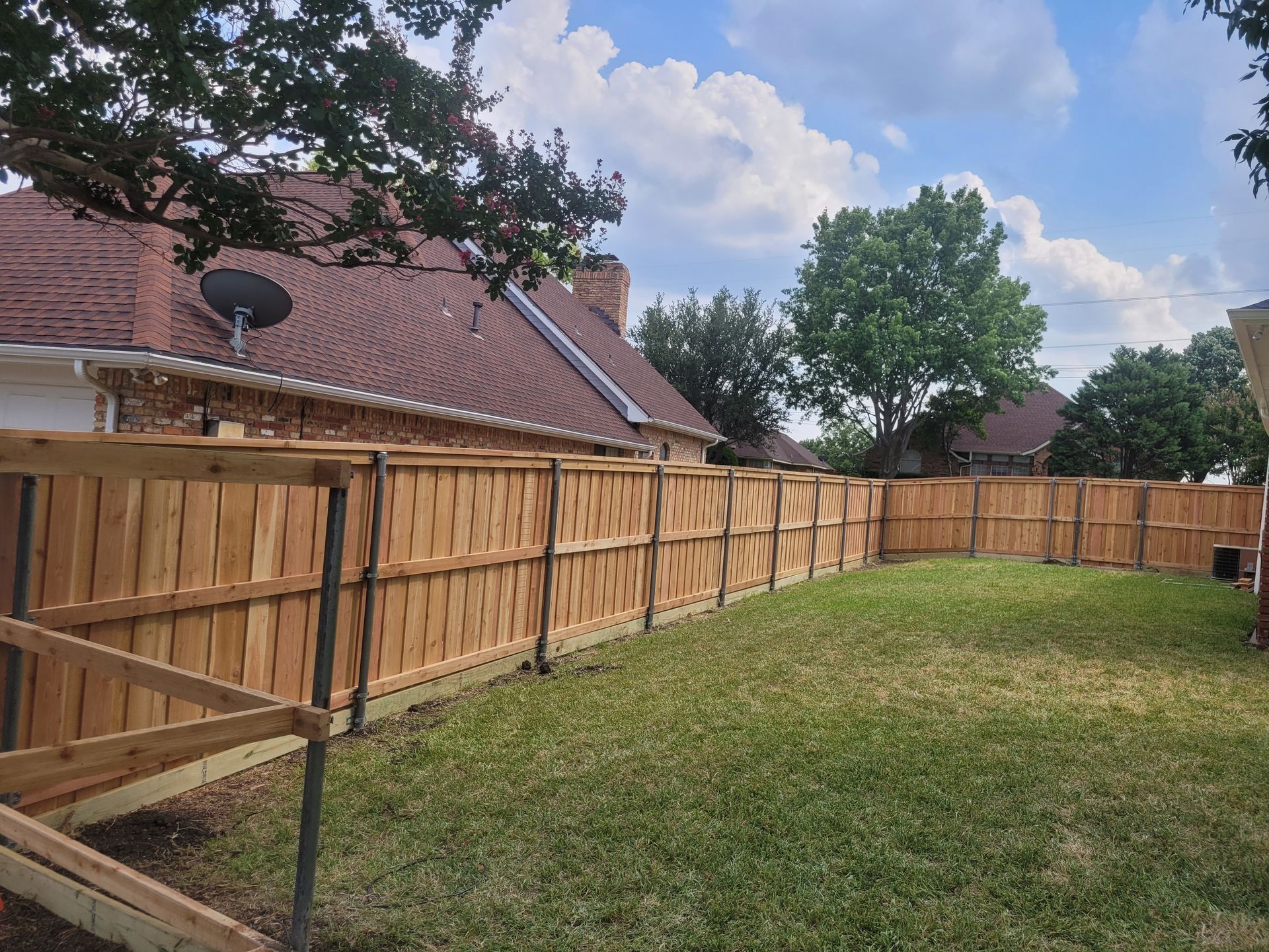 Backyard with a wooden fence, grassy lawn, and a red-roofed house under a partly cloudy sky