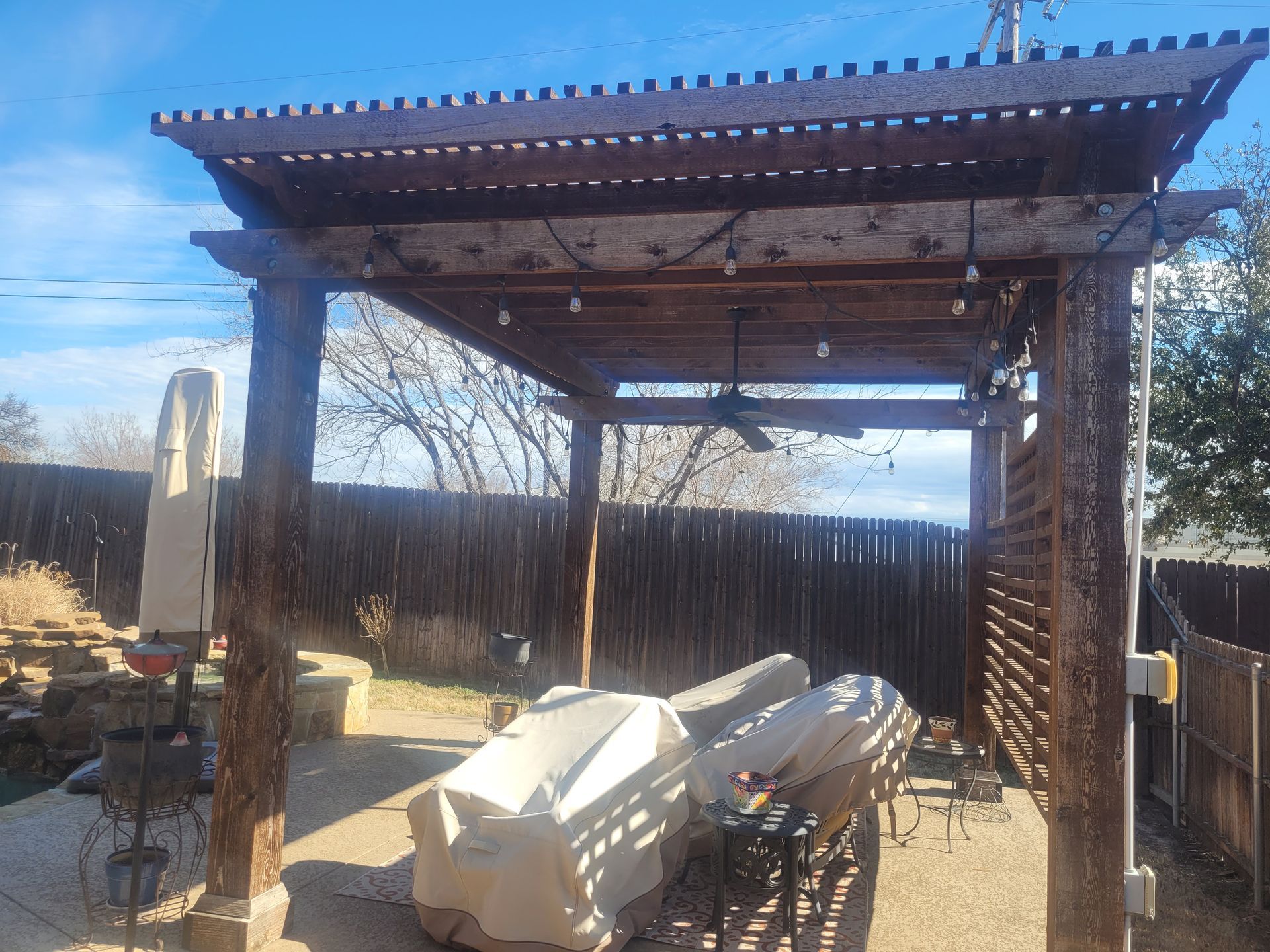 Outdoor pergola with wooden beams, covered furniture, and a bicycle in a fenced backyard under clear sky