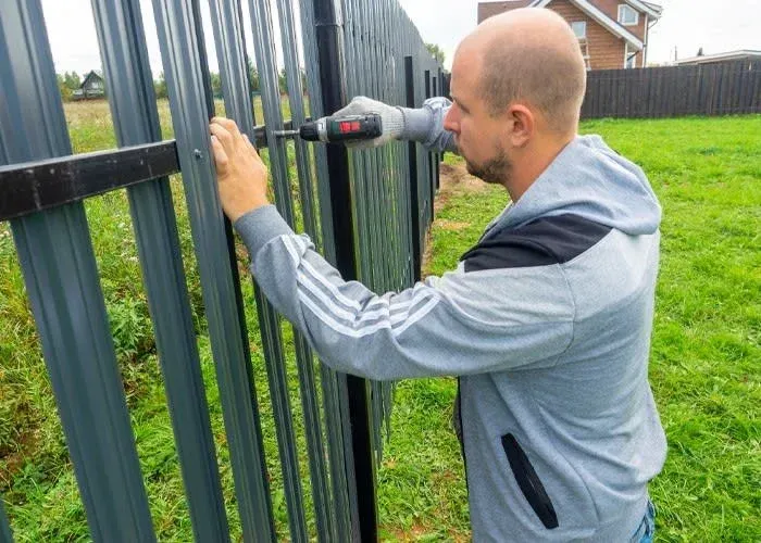 Man drilling a black metal fence outdoors beside a grassy yard