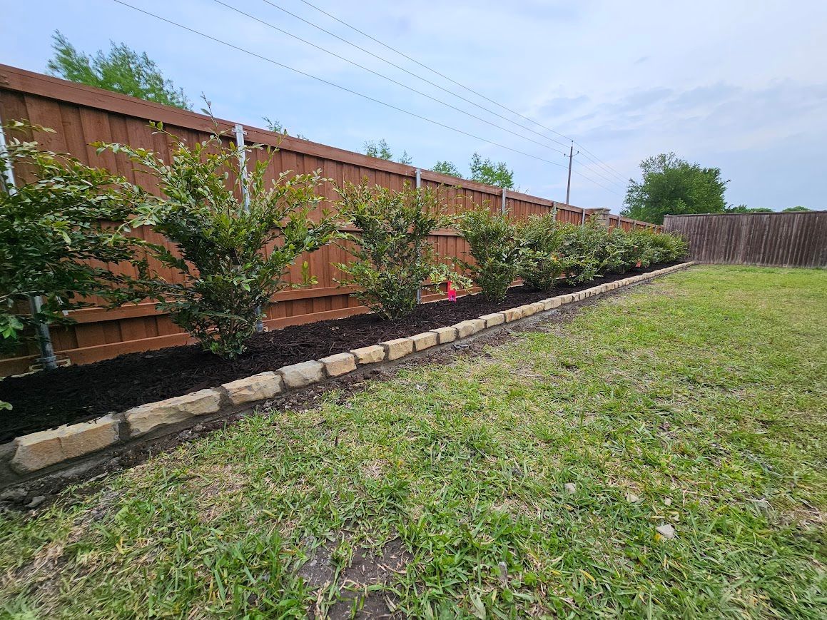 Grassy yard beside a wooden fence with a raised garden bed of shrubs and flowers under a cloudy sky