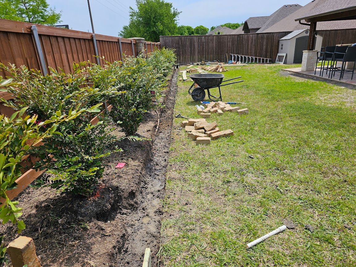 Backyard garden bed beside a wooden fence, with shrubs, grass, and a small wheelbarrow on the lawn
