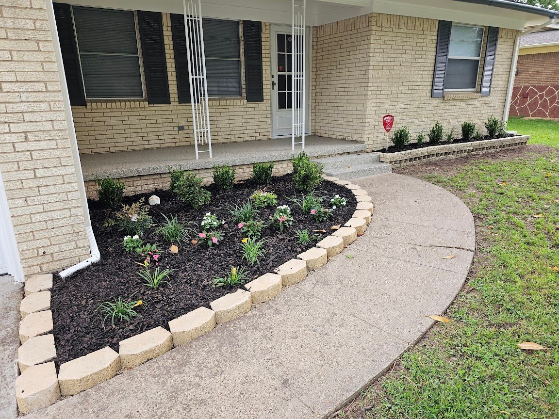 Curved concrete walkway beside a brick house, bordered by a raised flower bed with small shrubs and mulch.
