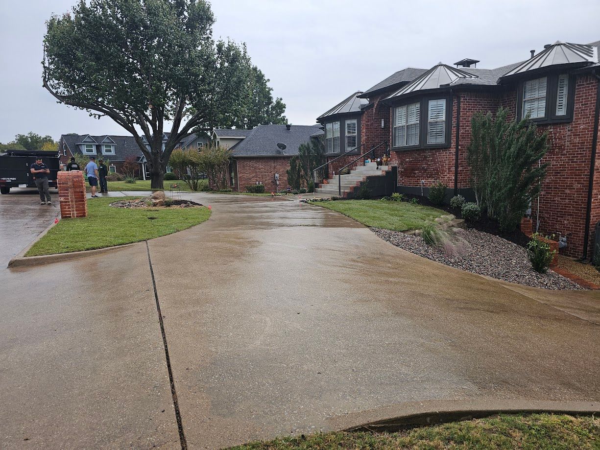 Wet suburban driveway beside red brick houses and a large tree on a rainy day