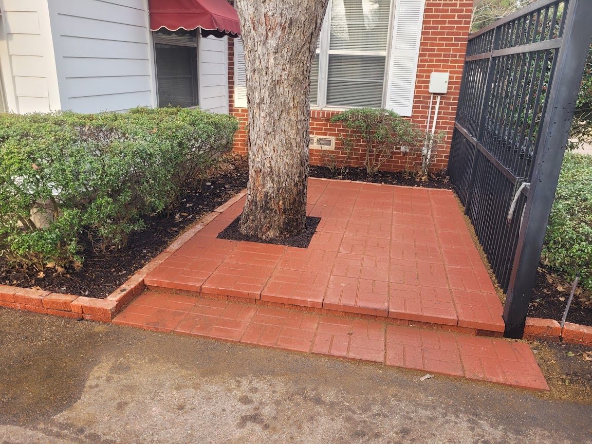 Brick patio around a tree beside a house, with hedges, red awning, and black metal fence.