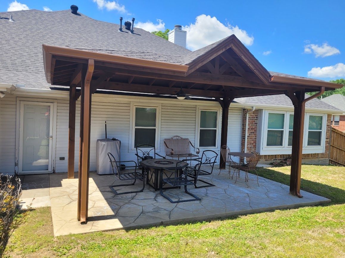Covered patio with outdoor dining table beside a white house under a shingled roof