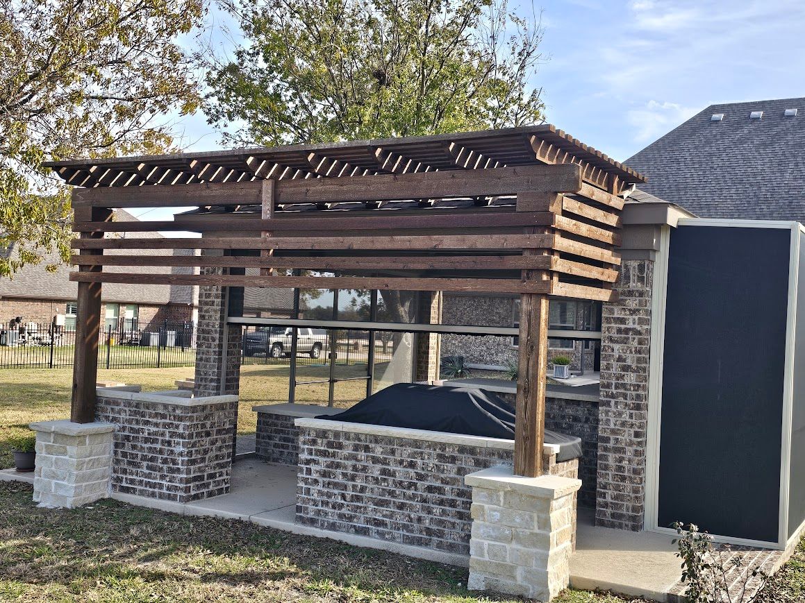 Wooden outdoor pergola over a stone bench beside a dark shed, in a grassy park area