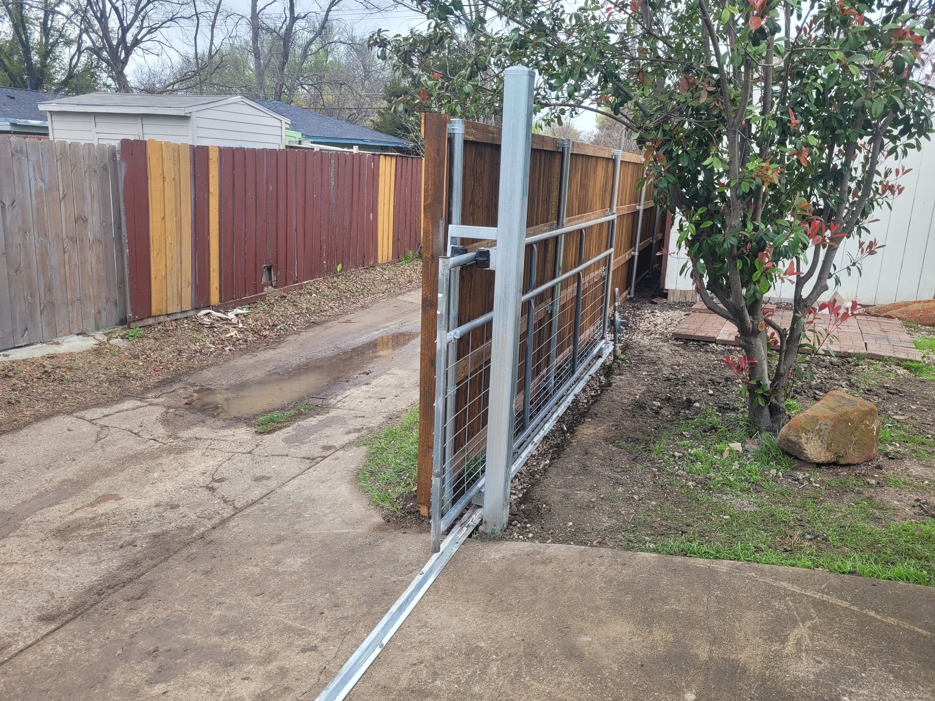Open metal gate beside a concrete driveway and wooden fence in a yard.