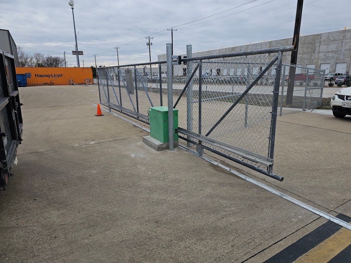 Chain-link gate beside a gravel lot with an orange traffic cone and gray industrial buildings