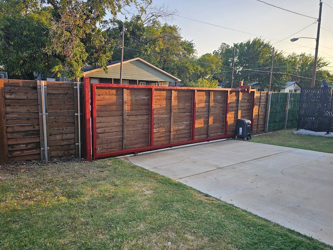 Red wooden fence and gate beside a driveway, with a grassy yard and trees in the background.