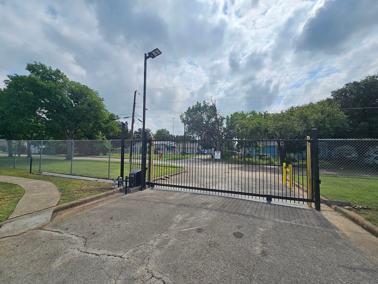 Chain-link fence gate at a park entrance beside a paved path and grass under cloudy skies