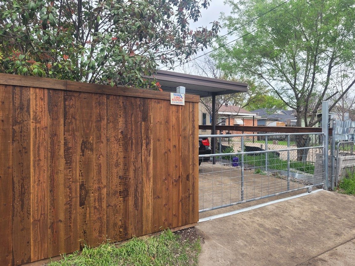Wooden fence beside a metal gate leading to a yard with trees and a small shed