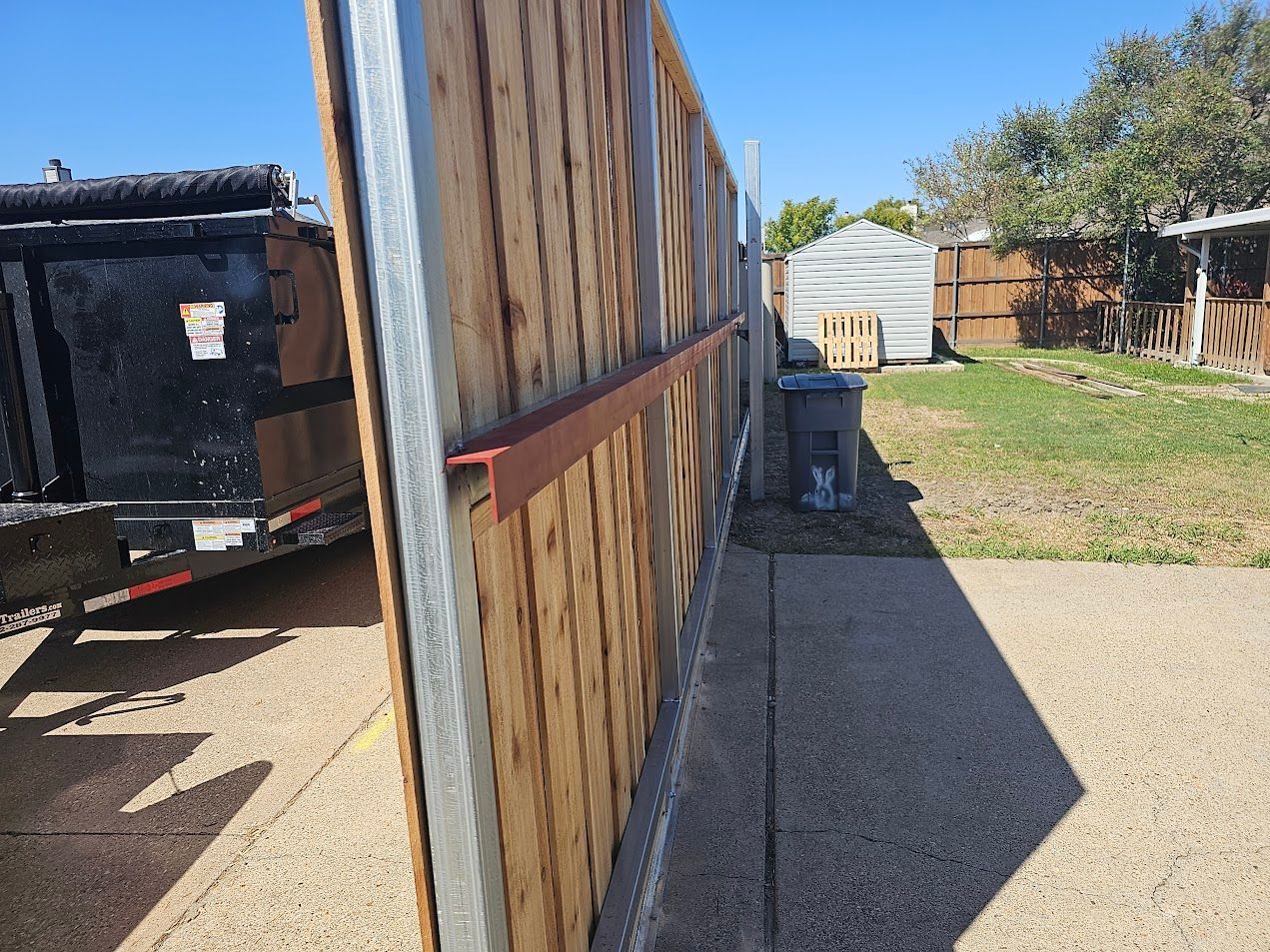 Wooden fence along a gravel driveway beside a trailer and yard under bright sunlight