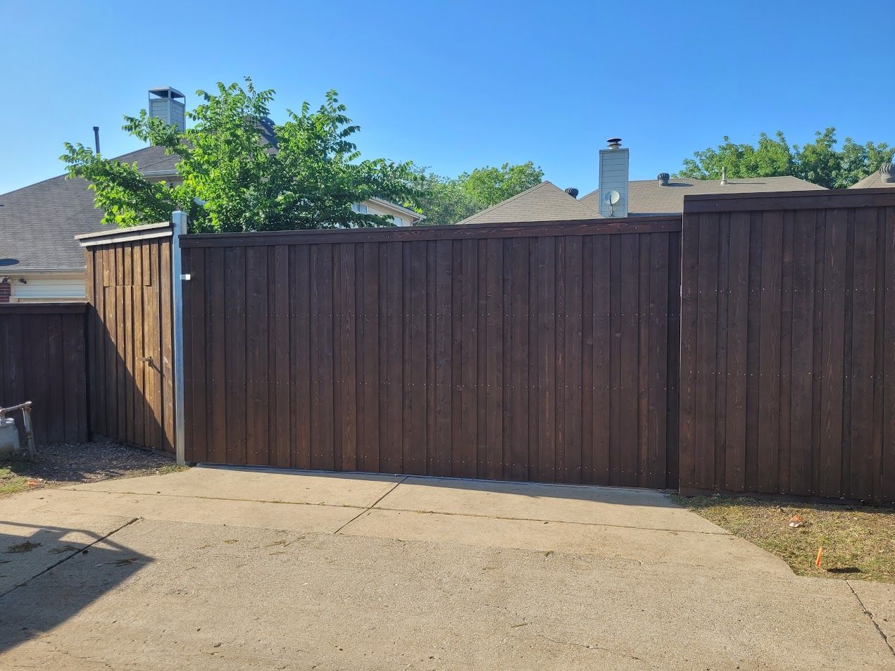 Brown wooden fence along a sandy yard with trees and blue sky behind it