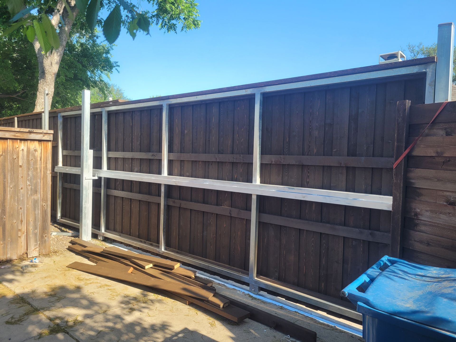 Wooden backyard fence under construction with horizontal braces, sunlit yard, and blue tarp on the right