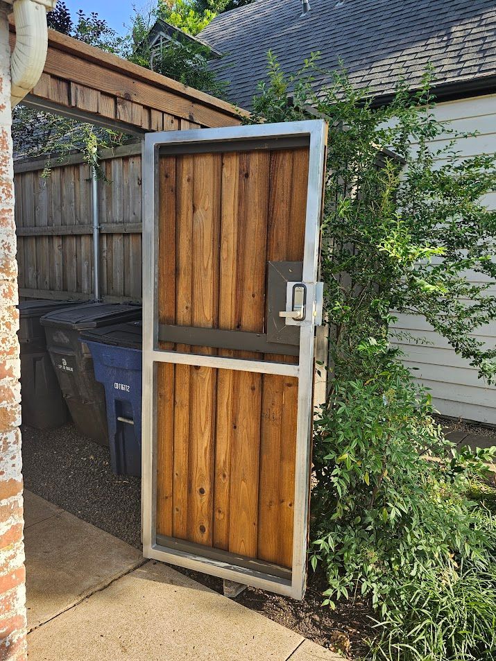 Open wooden gate with metal frame beside a driveway and trash bins in a backyard area