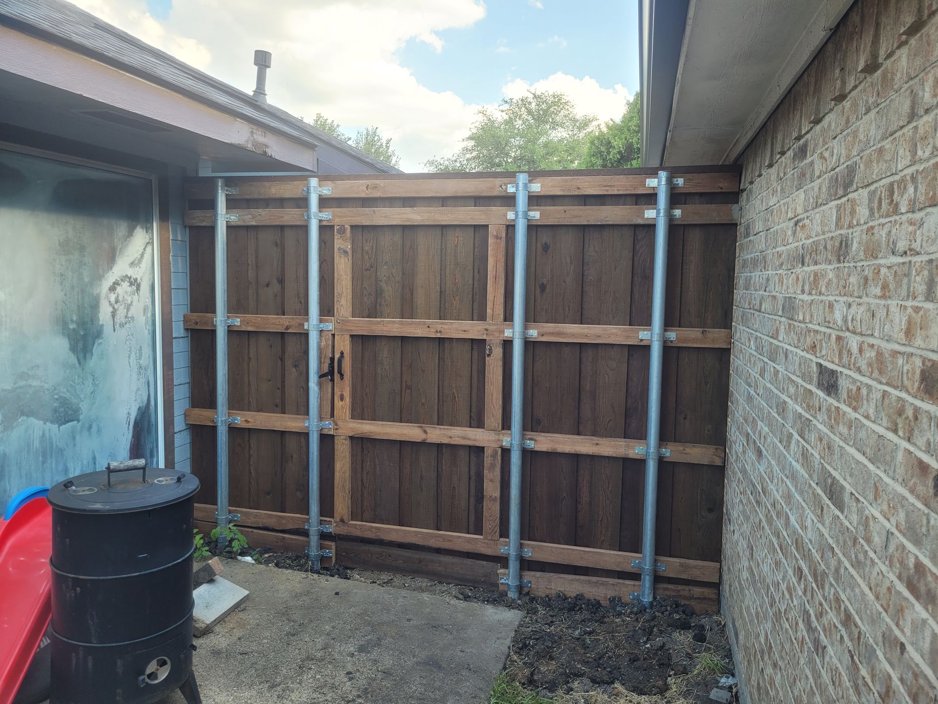 Backyard with a wooden fence panel between brick and stucco walls, beside a black barrel and red item