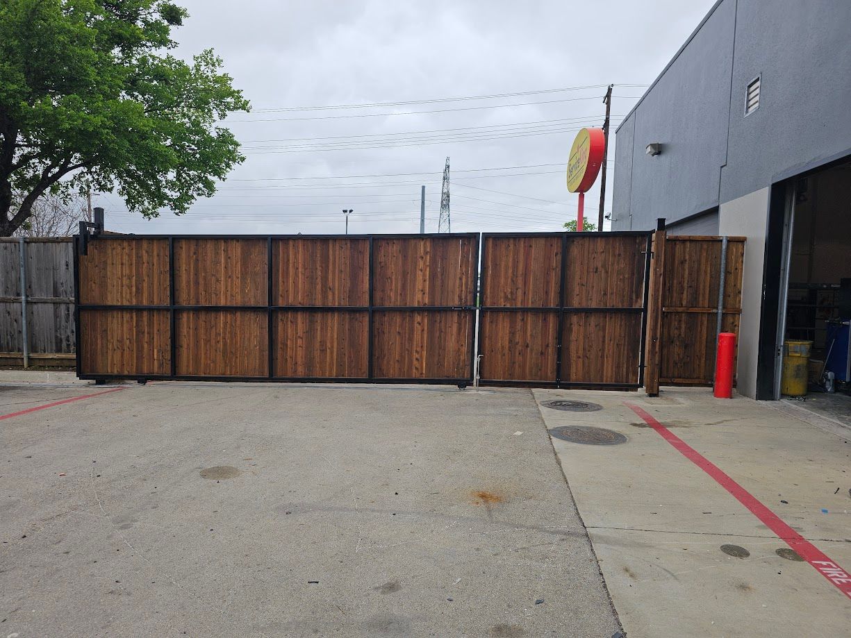Wooden gate blocking a paved driveway beside a gray building, with a tree on the left and a red curb line on the right.