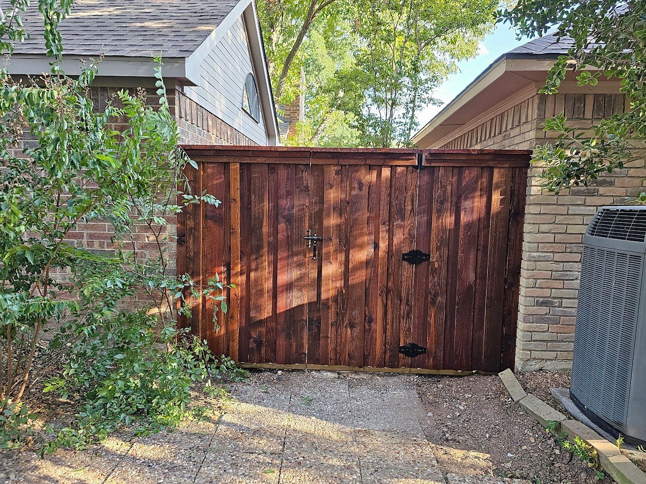 Wooden driveway gate between stone walls, with a leafy bush on the left and gravel ground.