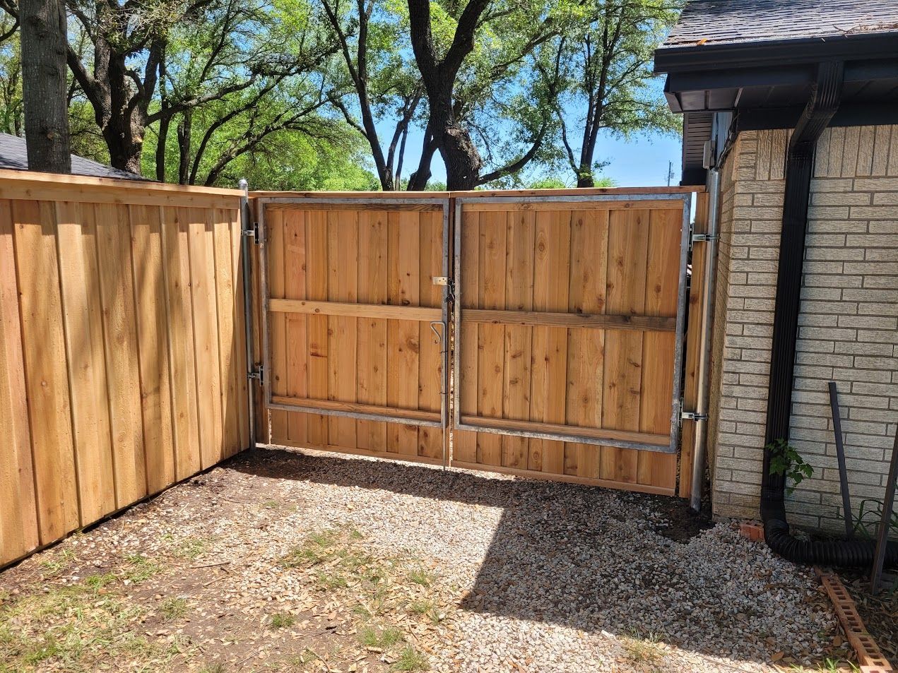 Wooden double gate between a fenced yard and a brick house under trees.