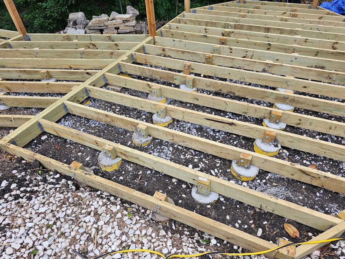 Wooden deck framing under construction on gravel, with joists and several support posts visible.