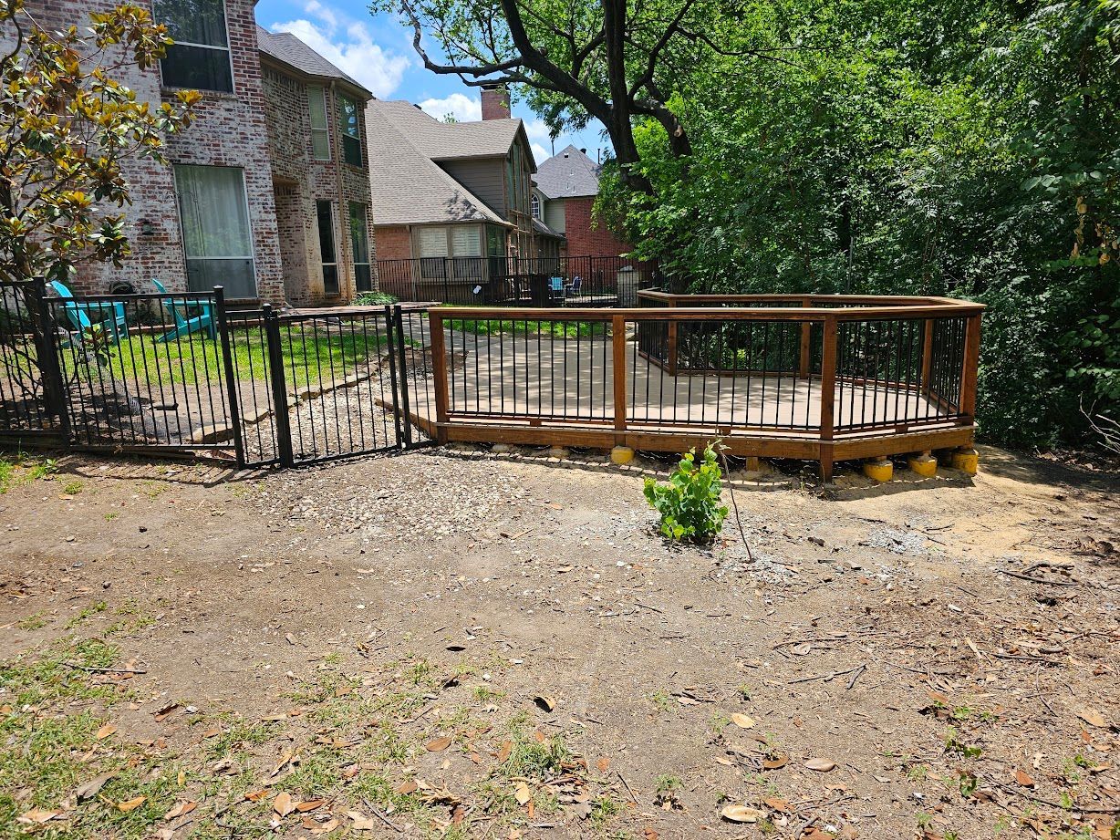 Backyard deck with wooden railing beside a stone house, surrounded by trees and dirt ground
