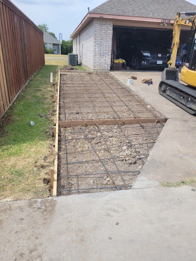 Driveway excavation for a new concrete slab beside a garage, with rebar grid and an excavator.