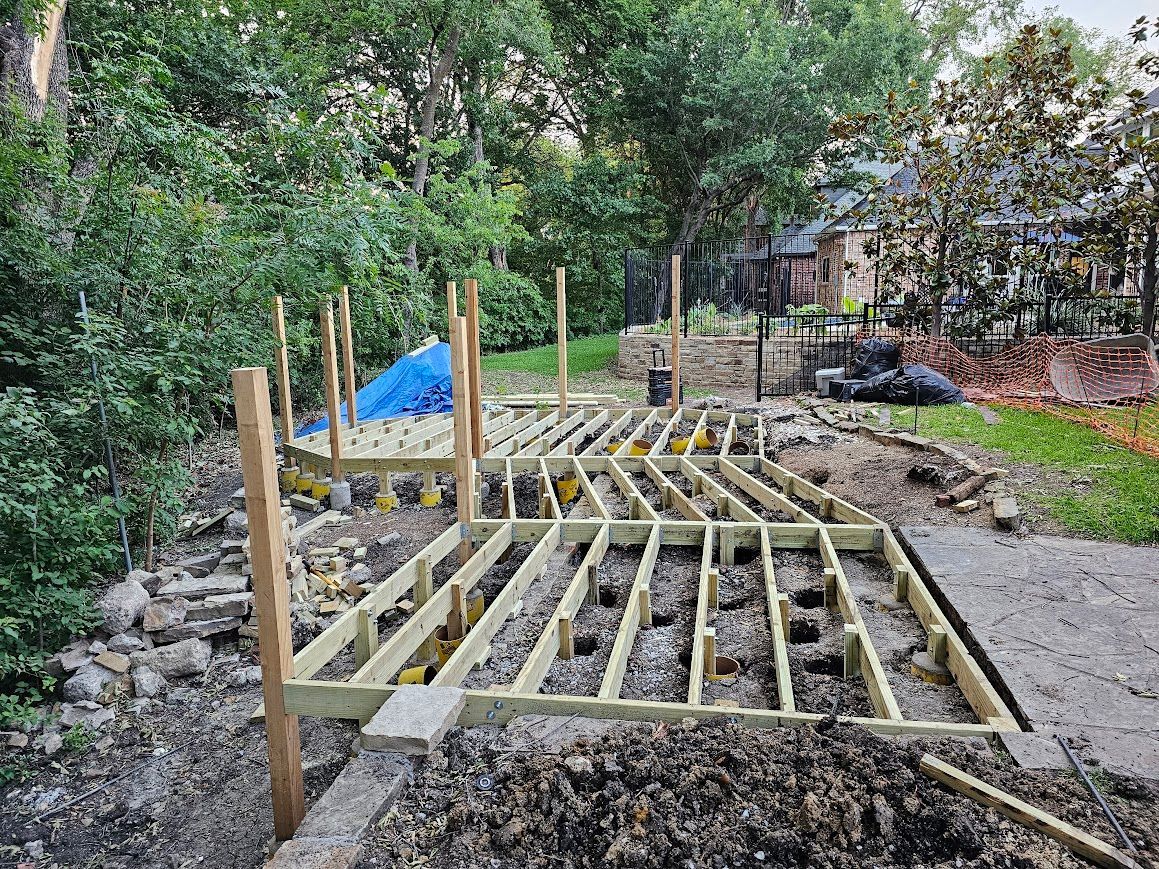 Wooden foundation frame under construction on a muddy site, with stakes and trees in the background.