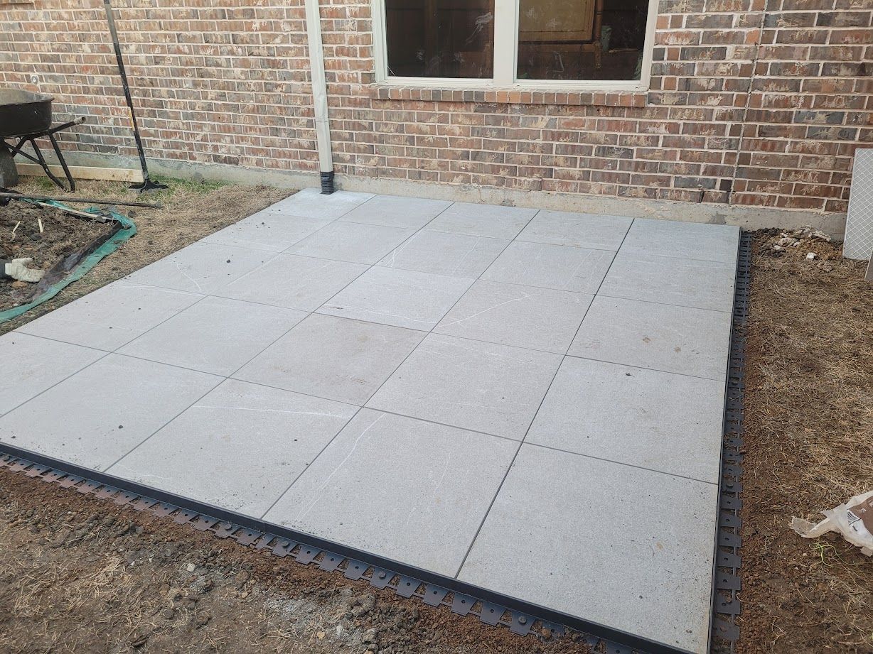 Concrete patio slab beside a brick house, bordered by gravel and soil