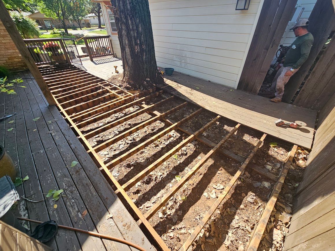 Deck framing under construction beside a tree and house, with exposed joists and debris.
