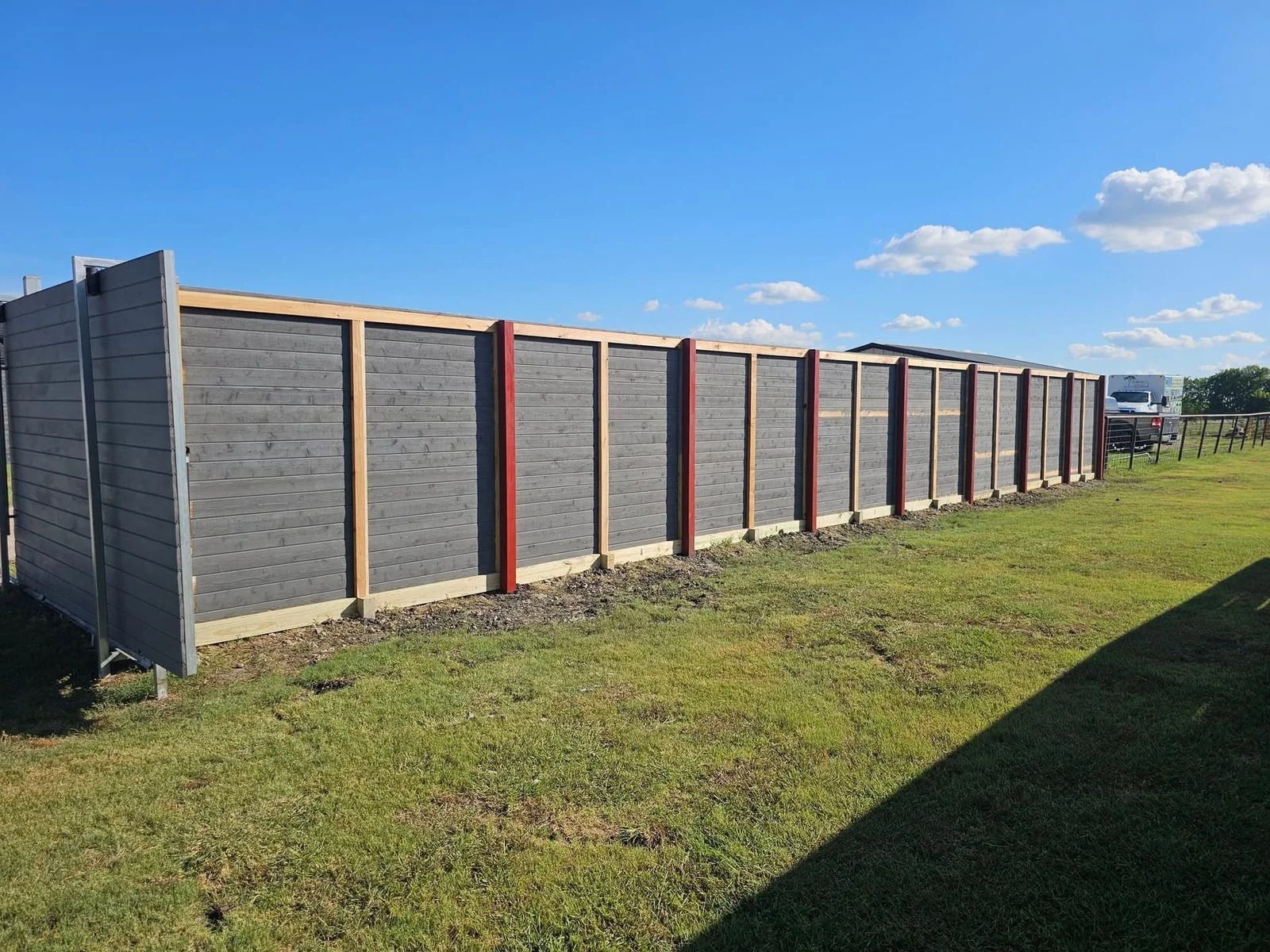 Long gray fence panels with a red post beside a grassy yard under a blue sky