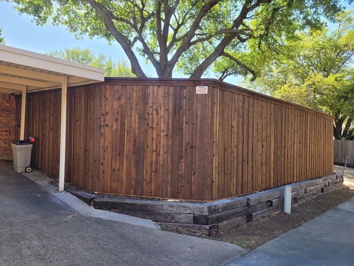 Wooden fenced storage area beside a driveway and carport, with trees overhead.