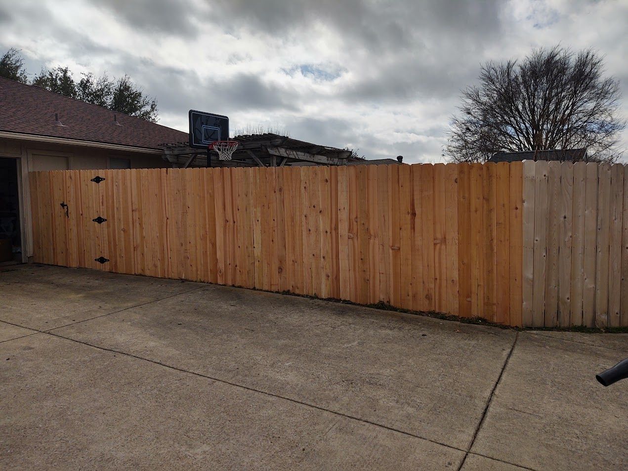 Wooden privacy fence along a driveway under a cloudy sky with houses and trees in the background