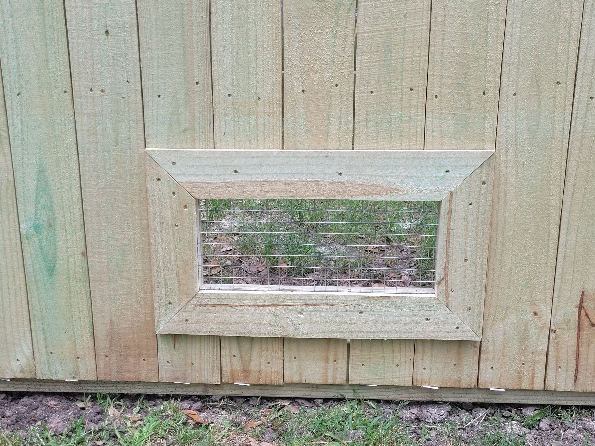 White pet door in a weathered wooden fence with a wire mesh opening.