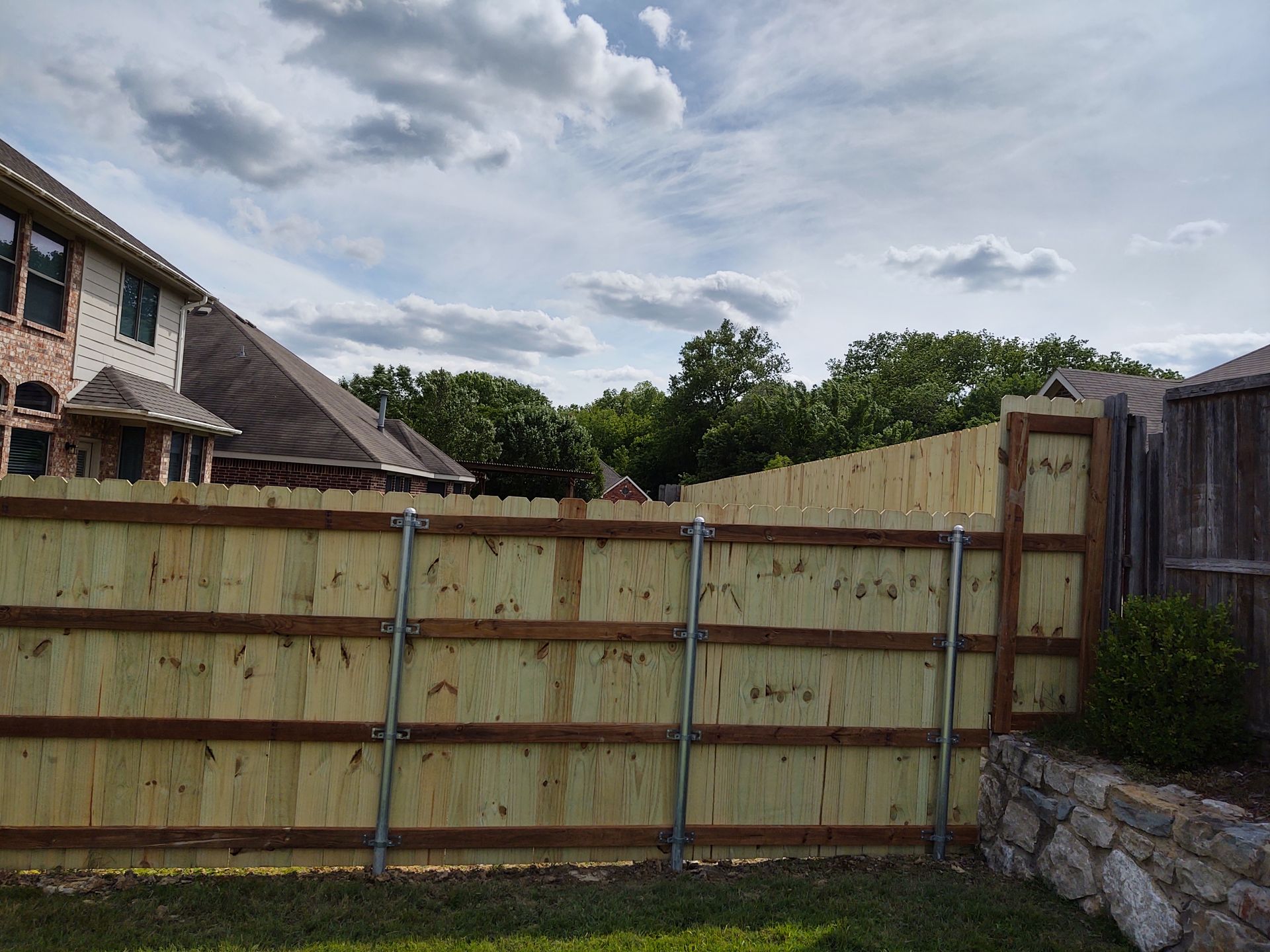 Wooden driveway gate with blue braces in front of a house and stone wall under a cloudy sky