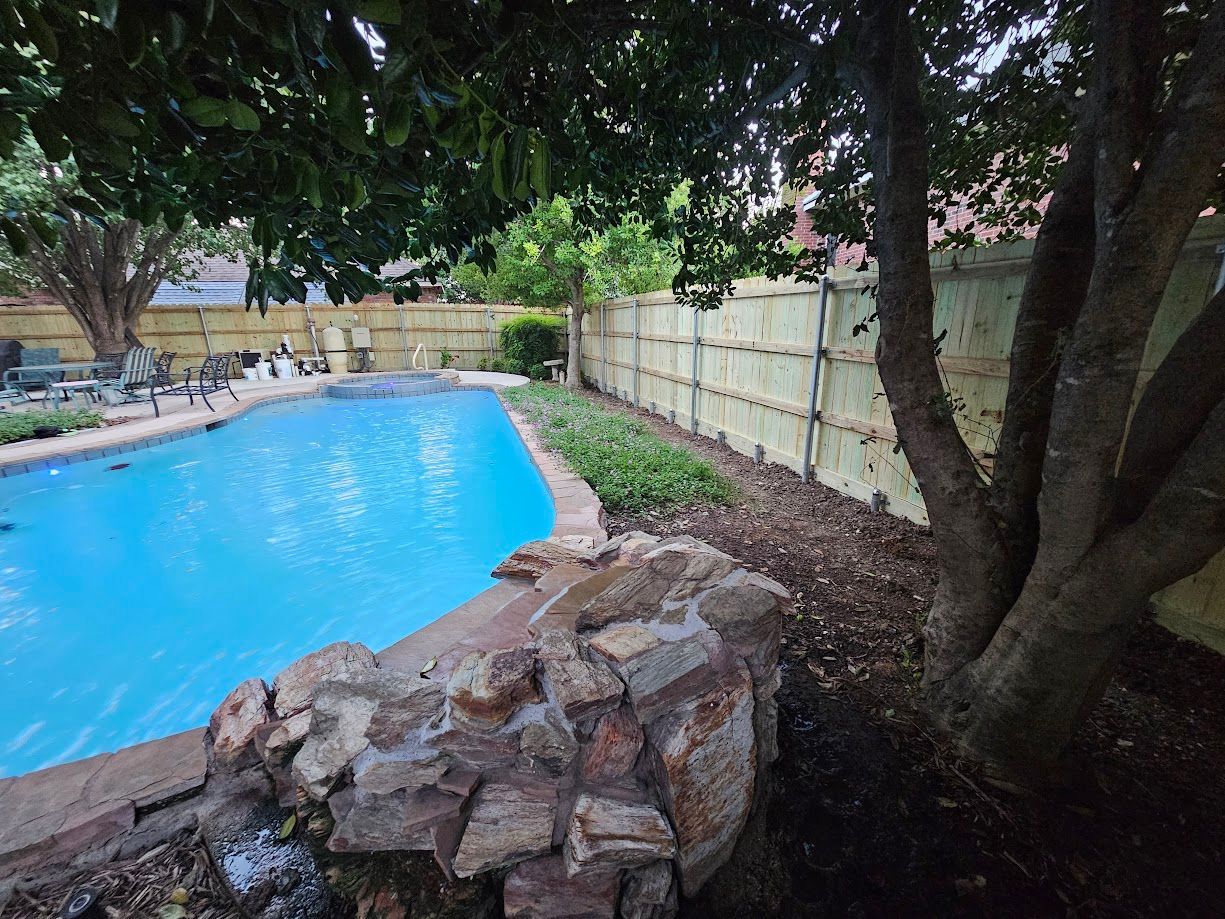 Backyard pool beside a stone planter, trees, and a white fence.
