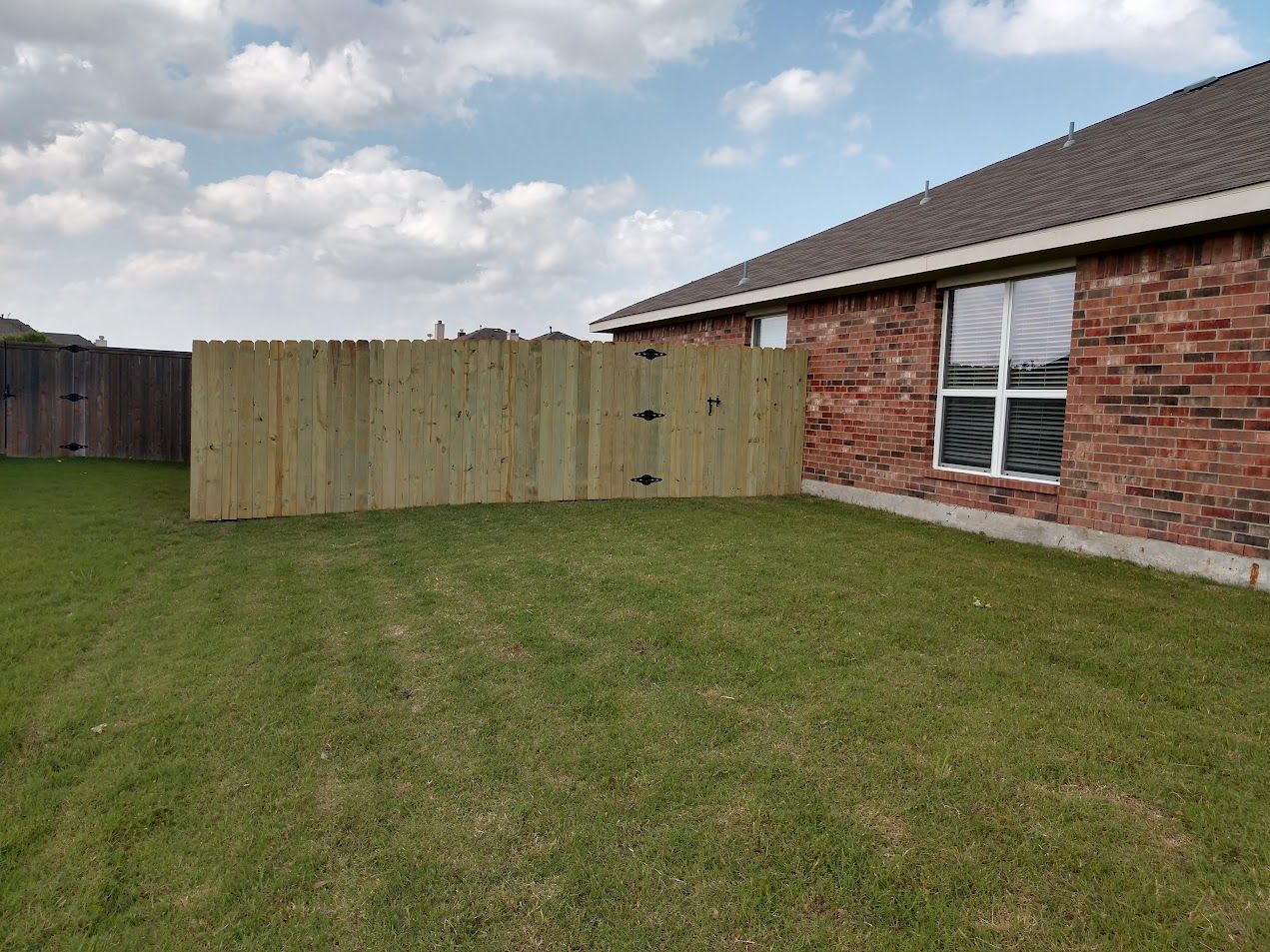 Backyard with green lawn, wooden fence, and brick house under a cloudy sky