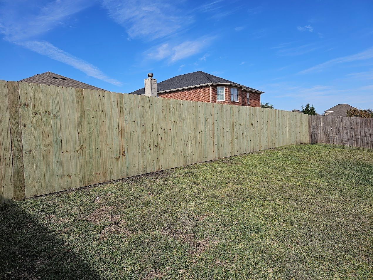 Weathered wooden fence beside a grassy yard under a clear blue sky, with houses in the background