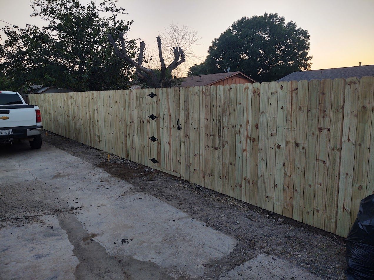 Wooden privacy fence beside a gravel driveway at dusk, with parked vehicles and trees in the background