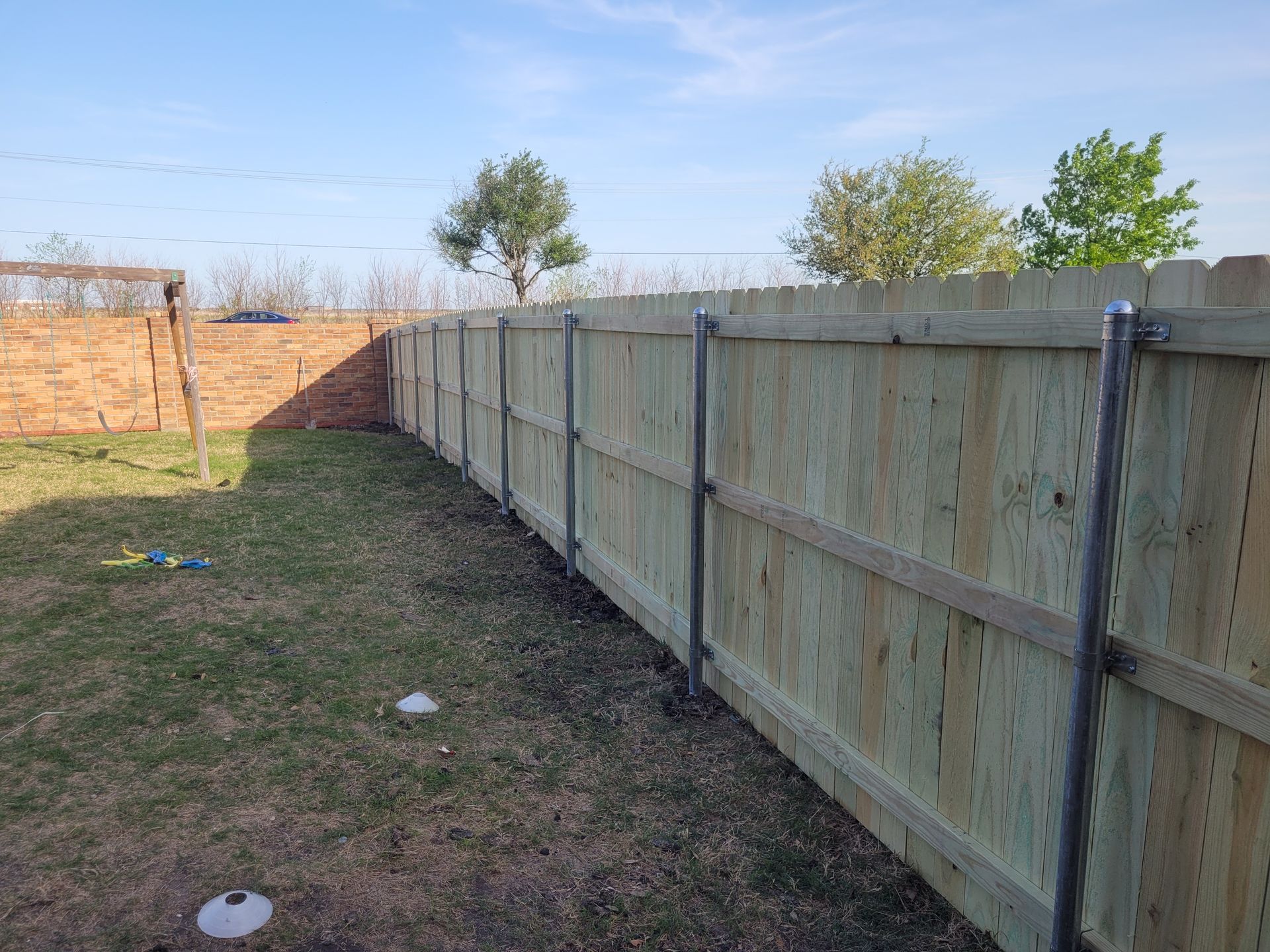 Long wooden privacy fence along a narrow grassy yard with brick wall at the far end