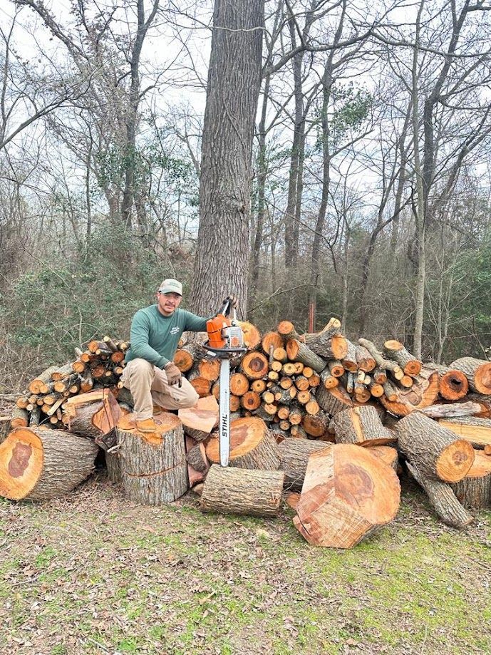 Two people with a chainsaw posing beside a large pile of cut logs in a wooded area.
