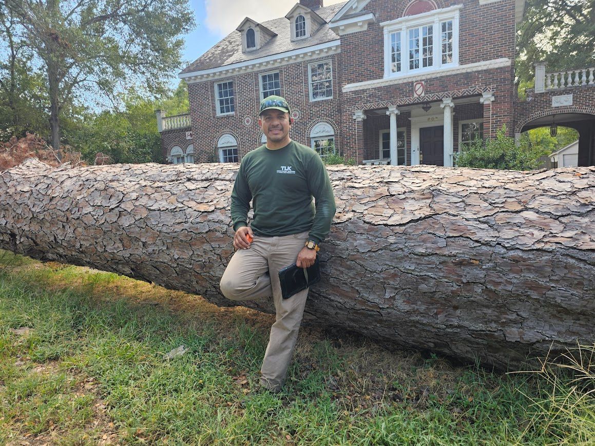 Man posing beside a massive fallen tree trunk in front of a brick house and yard.