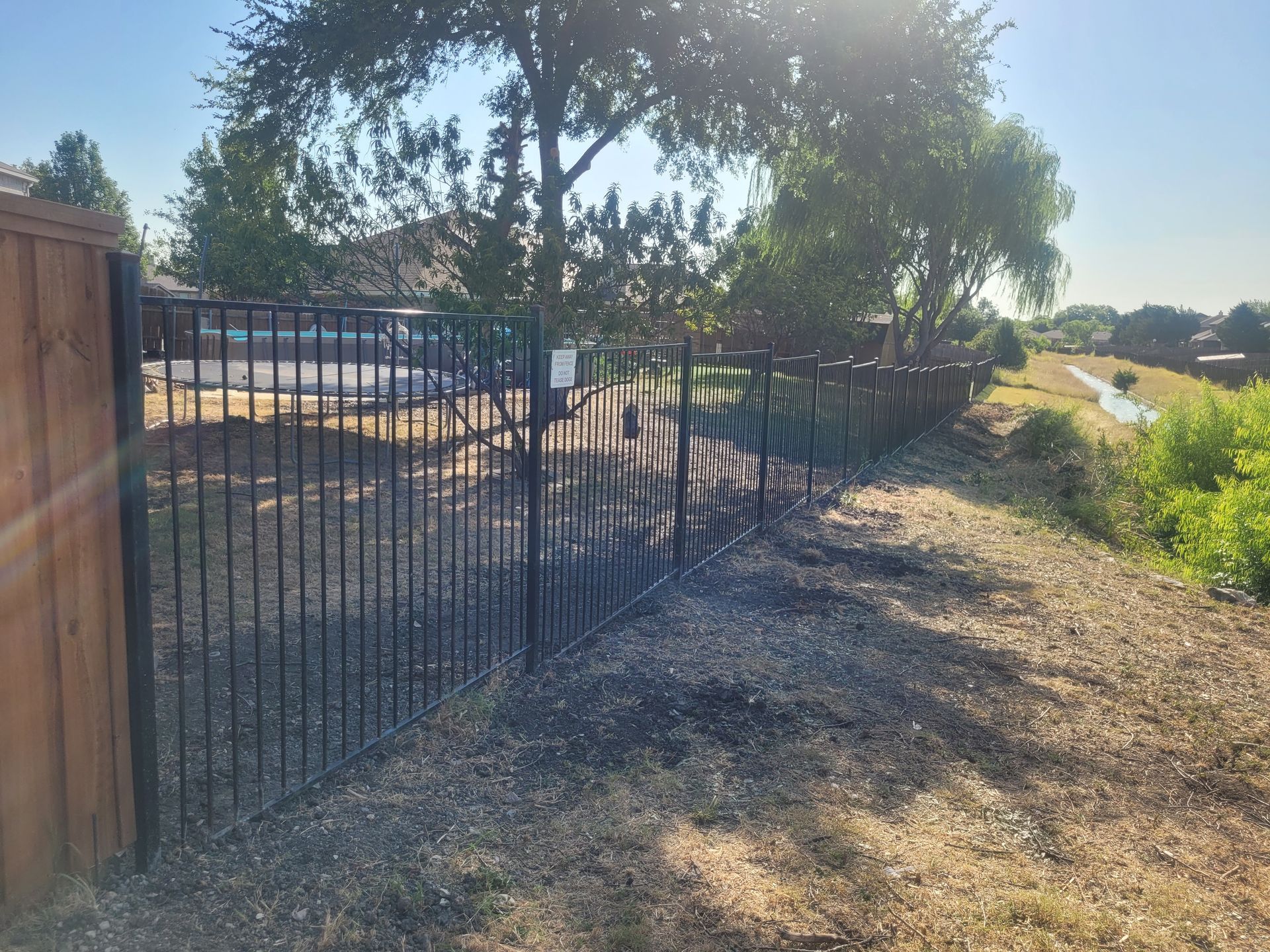 Sunlit fenced dirt path beside a creek, with trees and a wooden gate in the foreground.