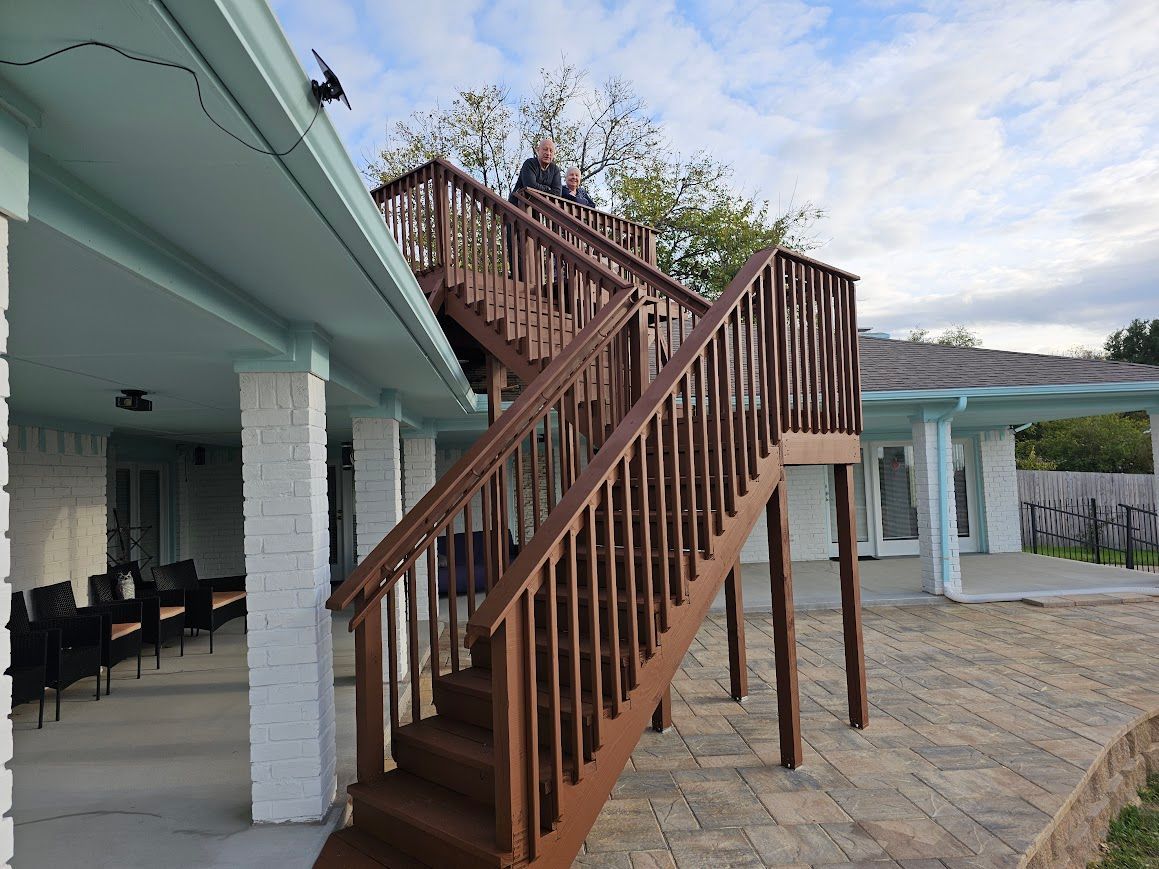Outdoor wooden staircase leading to an upper deck beside a white house and stone patio
