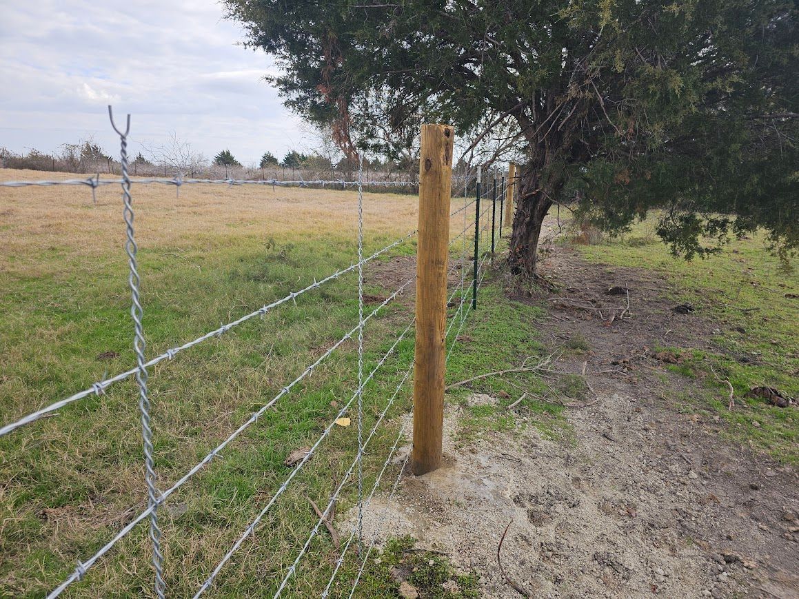 Wire fence and wooden post beside a dirt path under a tree in a rural field