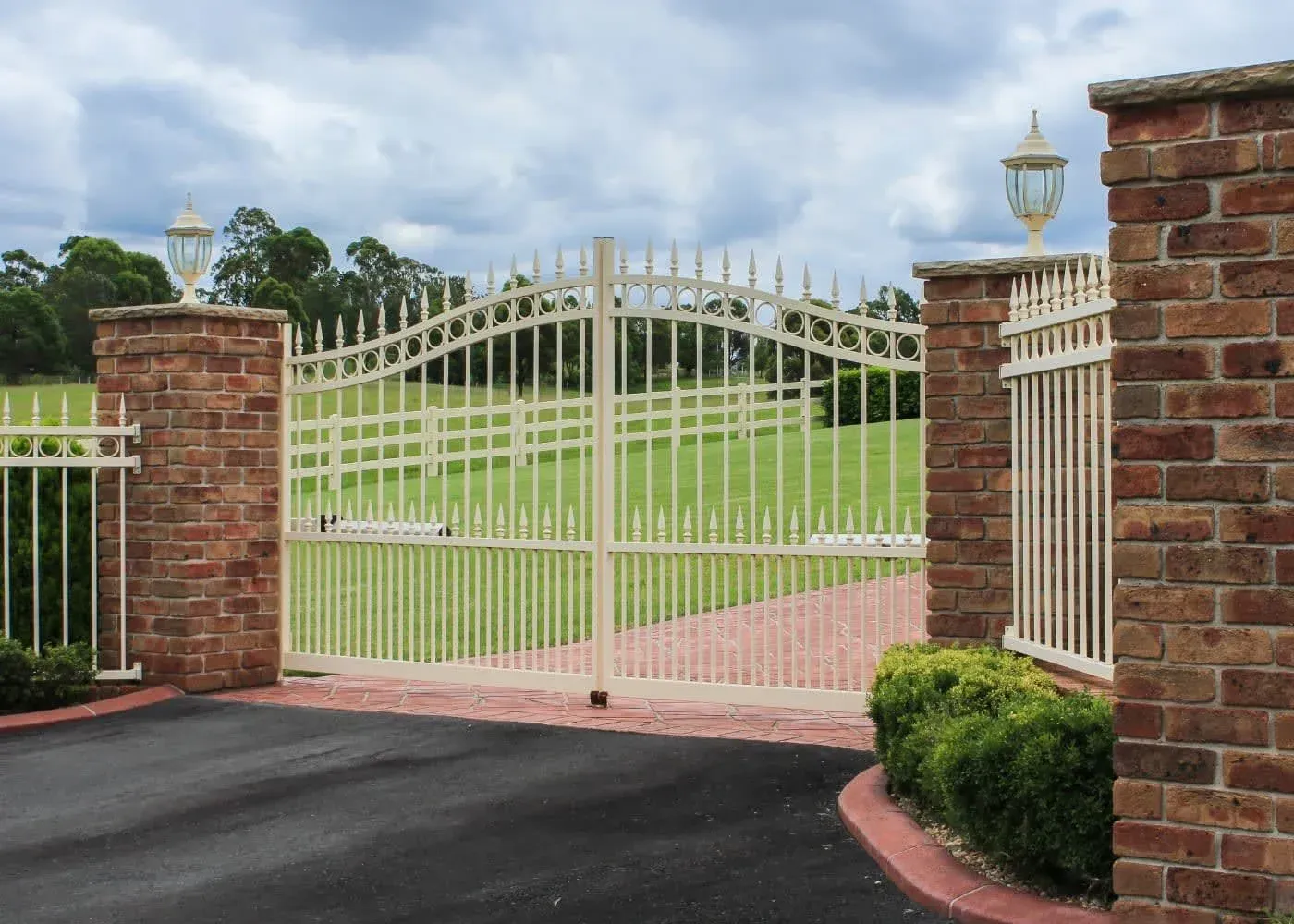 Cream-colored wrought-iron gate between brick pillars, opening to a landscaped driveway and lawn.
