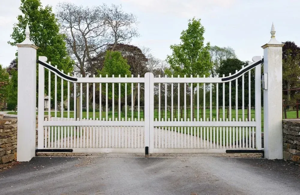 White metal driveway gate between stone pillars, closed across a paved entrance with trees beyond.