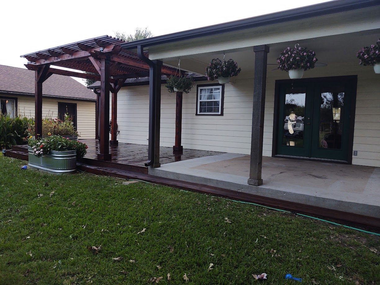 Covered walkway outside a beige building with dark doors, potted flowers, and green lawn along the front