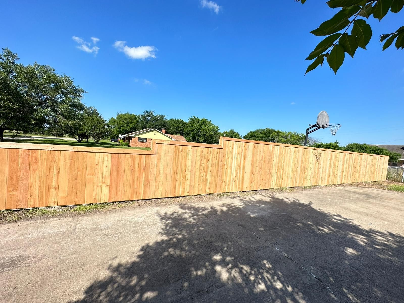 Wooden fence along a paved driveway under a clear blue sky, with trees and a basketball hoop nearby
